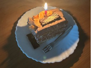 完成したチョコバナナケーキをフォークとともに皿に盛りつけ、ケーキの上には網チョコとスライスアーモンドをトッピングし、火のついたロウソクを立てています。