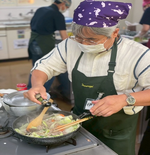 男性が、野菜と肉を炒めたフライパンに調味料を加えて味付けをしています。