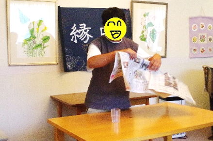 一人のメンバーが新聞紙を使ってコップを消す手品を披露しています。