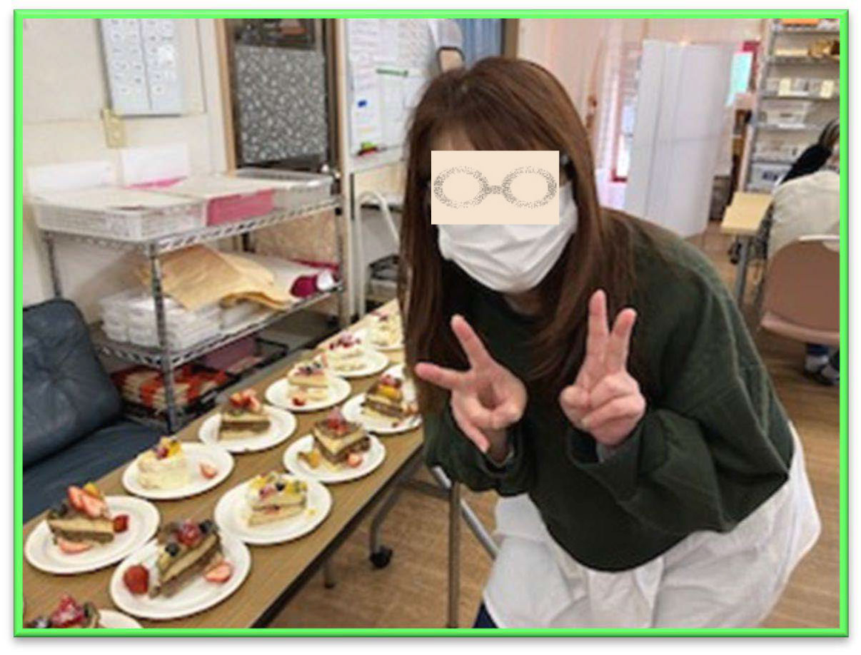 所長が、ケーキが置かれた机の横でピースサインをしている。