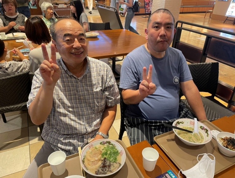 男性2人が横並びで昼食のラーメンを食べながら、ピースサインをして写真に写っています。